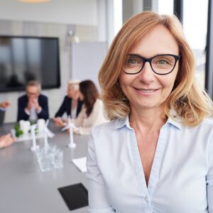 Smaidīga, pārliecināta vadītāja brillēs un gaišā kreklā uz sapulču telpas fona, kurā redzama komanda un vēja ģeneratoru modeļi.