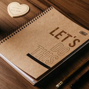 Spiralveida piezīmju grāmatiņa ar brūniem kraftpapīra vākiem un uzrakstu "LET'S DO THIS" guļ uz koka galda blakus zīmulim un sirds formas koka dekors.