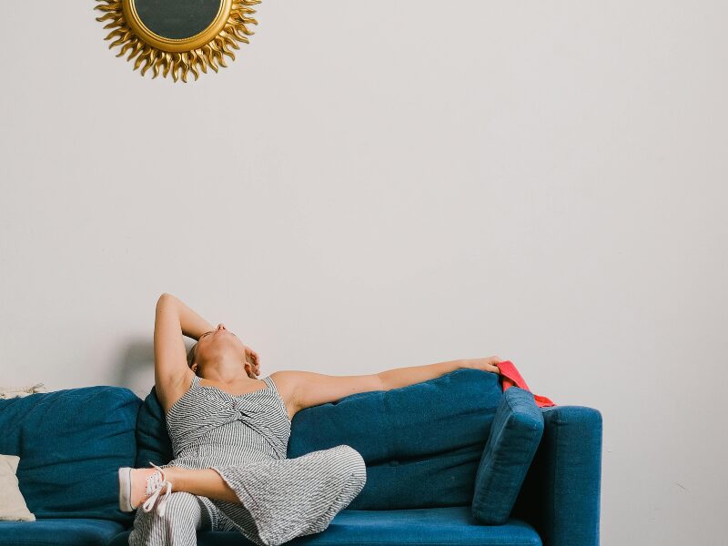 Sieviete atslābusi guļ uz zilas dīvāna malas, atgāzusi galvu atpakaļ, telpā ar gaišu sienu un dekoratīvu spoguli virs dīvāna, radot noguruma un atelpas sajūtu.
