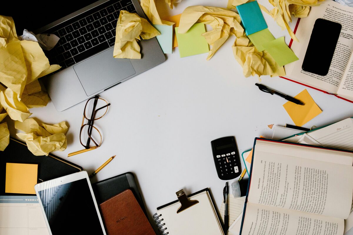 Darba galds ar izkliedētu nekārtību – papīri, uzlīmes, portatīvais dators, kalkulators un biroja piederumi.