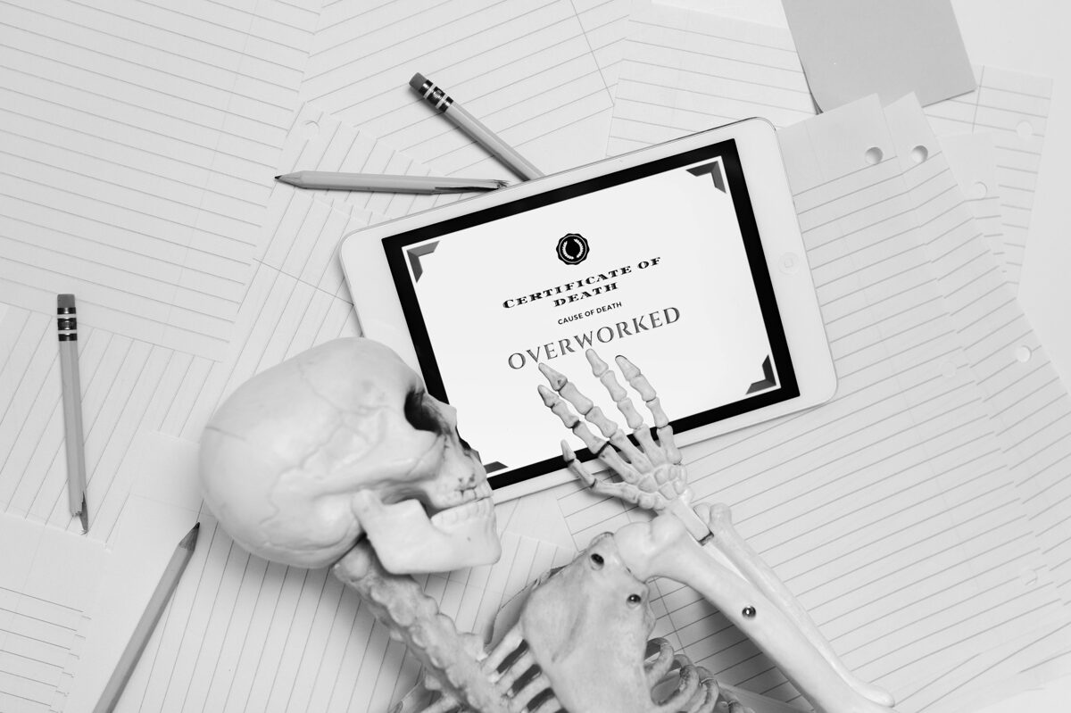 Attēlā redzams skelets, kas guļ uz papīru un zīmuļu kaudzes, blakus planšetdators ar uzrakstu “Certificate of Death – Cause of Death: Overworked”. Tas simbolizē pārmērīgu izdegšanu darbā un ironiski ataino sekas, ja cilvēks ilgstoši ignorē savas robežas un nepievērš uzmanību emocionālai pārslodzei.